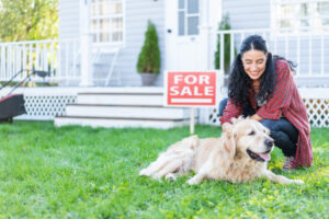 Smiling woman in front of "For Sale" sign, kneeling next to golden retriever