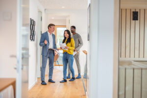An adult caucasian male real estate agent giving an apartment tour to a young diverse couple. The real estate agent is wearing a fancy suit while the couple is wearing casual clothes. Together they are walking through the hall of the apartment. The rooms have modern decor with wooden details.